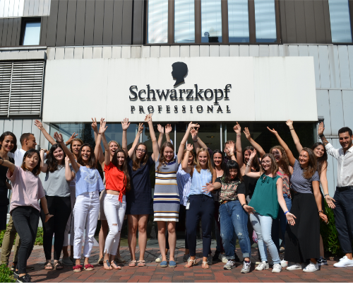 فريق يضم مجموعة متنوعة من موظفي Henkel يشجّعون أمام مبنى Schwarzkopf الاحترافي ويرفعون أياديهم