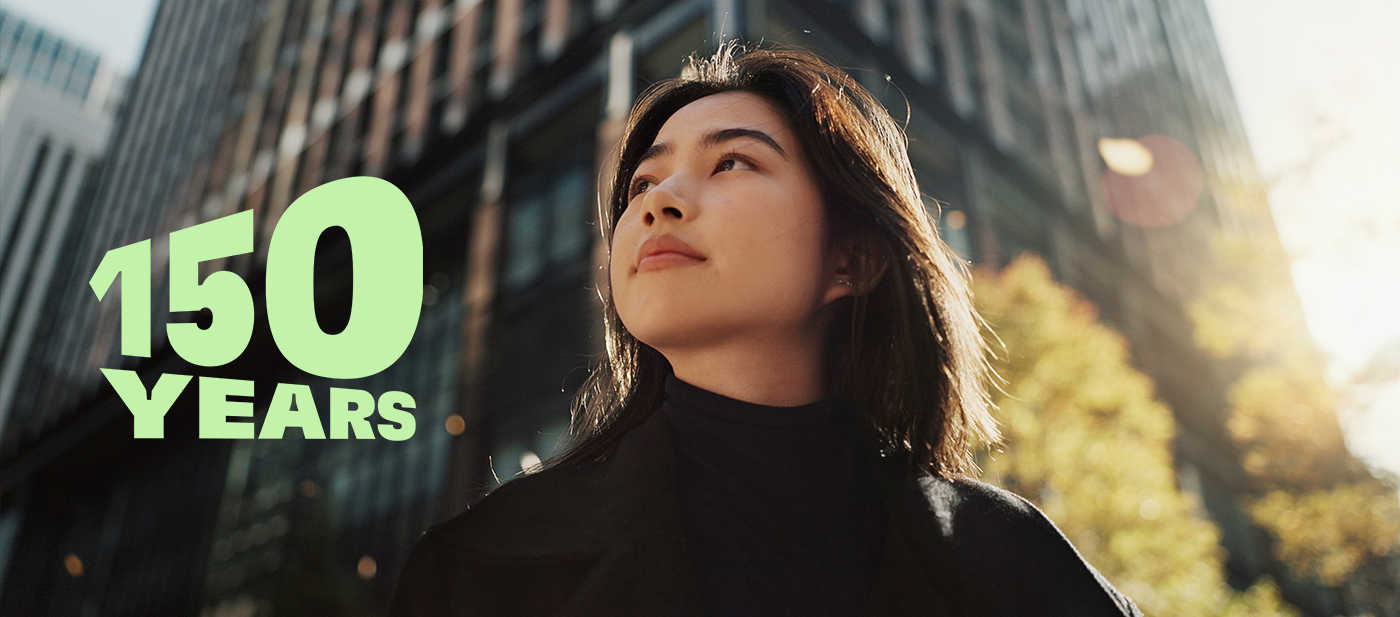 Grøn tekst med teksten »150 ÅR« over et moderne bybillede med høje glasbygninger og efterårstræer, der symboliserer 150-års jubilæet.
