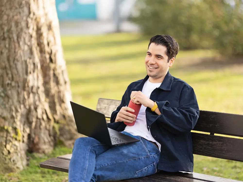 En Henkel-medarbejder sidder på en bænk i en park og arbejder på sin bærbare computer. 