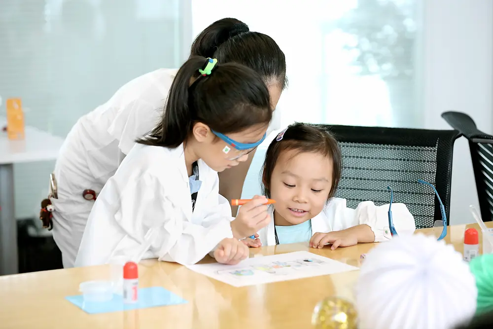 To børn og en kvinde i en forskerfrakke farver et billede ved et bord