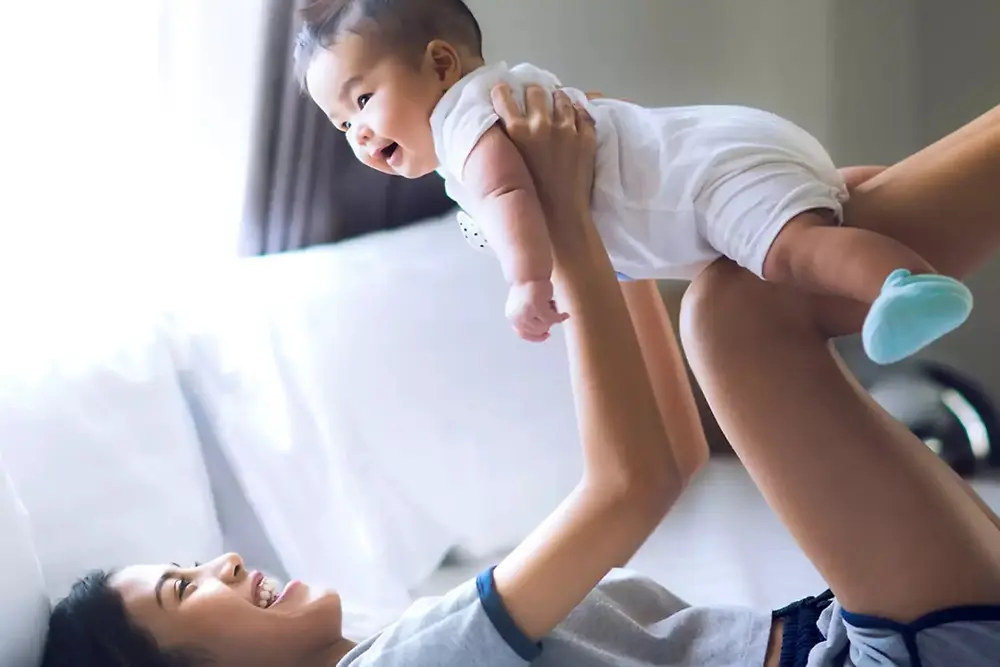 Mor løfter og leger med en smilende baby indendørs i et lyst hjemmemiljø.
