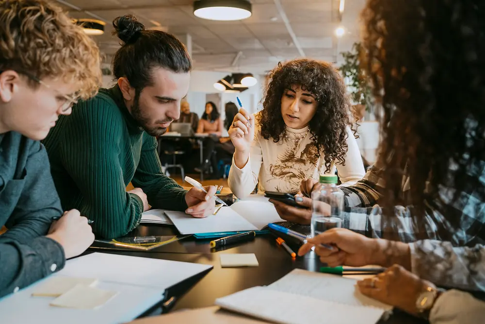 En gruppe mennesker sidder samlet ved et bord i et moderne indendørs kontorlandskab og arbejder sammen på dokumenter, notesbøger og digitale enheder. Der ligger forskellige kuglepenne, papirer og post-it-sedler spredt ud over bordet, mens gruppen diskuterer og tager noter.