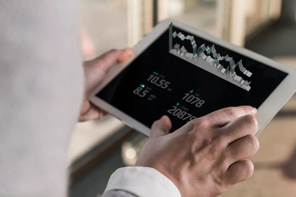 En person, der holder en tablet med digitale kort og taldata, set over skulderen i et lyst indendørs miljø.